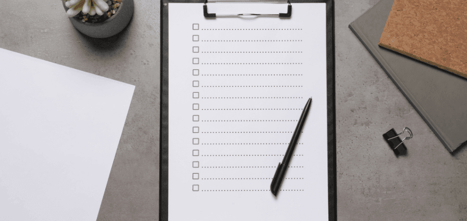 A blank checklist on a clipboard with a black pen resting on it, placed on a gray desk with a few office supplies around. This image represents the importance of standardized inspection checklists for thorough facility evaluations.